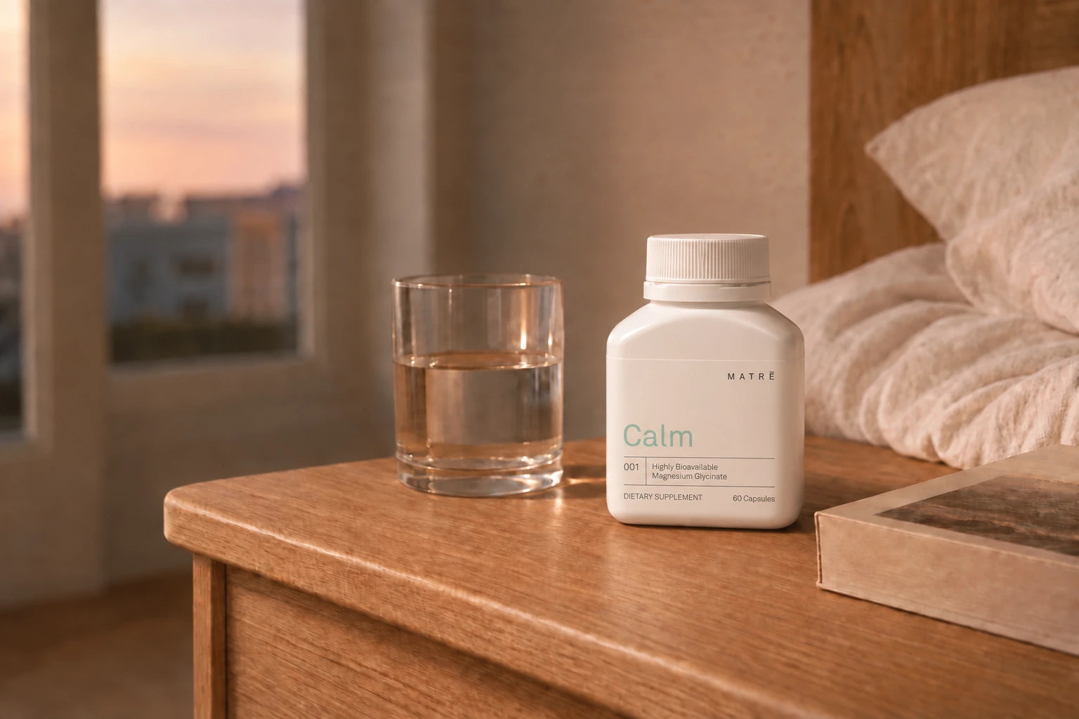 Matrē Calm bottle beside water and a book on a pale oak bedside table, with a green plant in the background